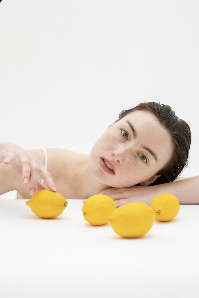 Een vrouw die op een witte ondergrond ligt achter vier verse citroenen, wat frisheid en natuurlijke vitamine C suggereert.